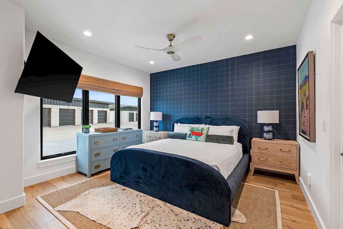 A modern bedroom with a navy blue accent wall, king bed, matching blue and wood furniture, a wall-mounted TV, large window, and cozy rug.
