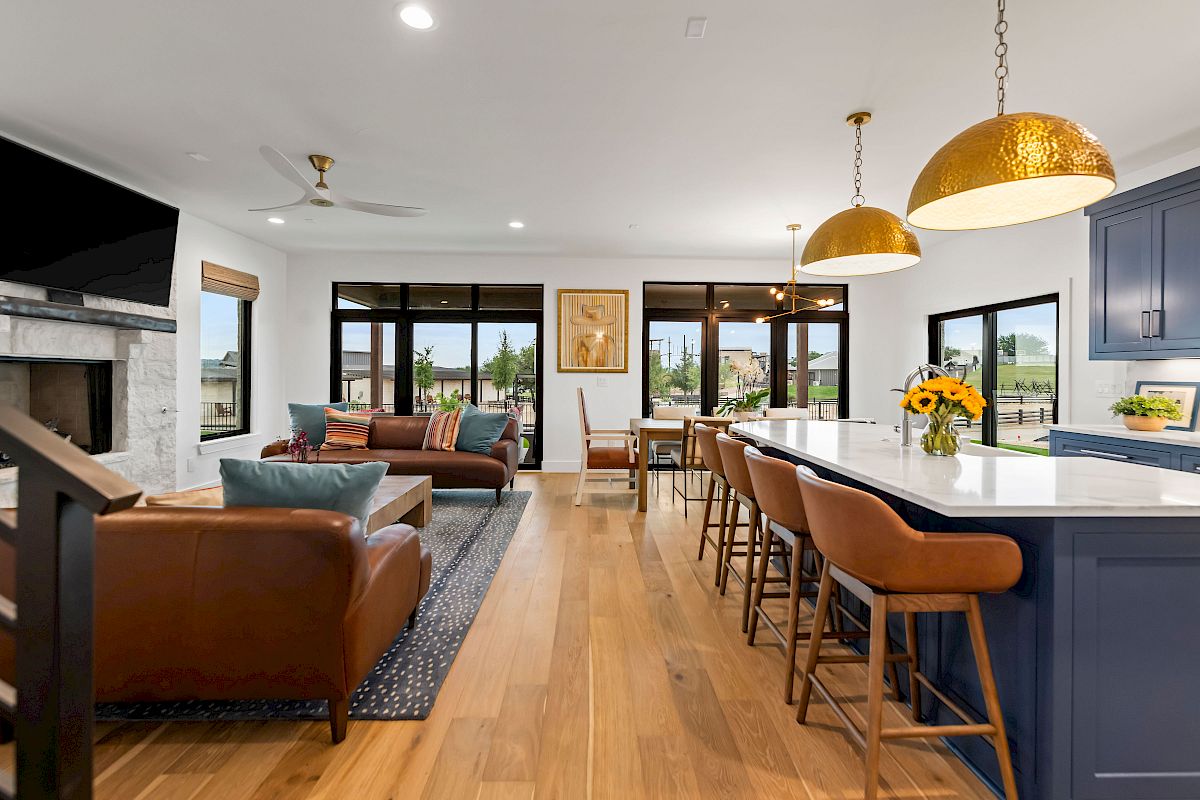 A bright, open living area with a kitchen island, blue cabinets, tan bar stools, leather couches, a fireplace, and large windows.