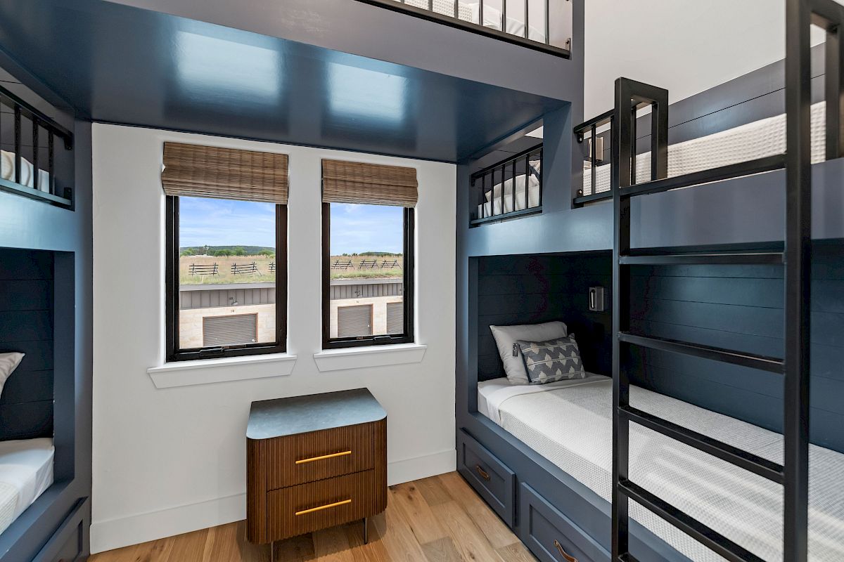 A blue-themed dorm with four bunk beds, wooden drawers, and two windows, plus a small nightstand under the windows.