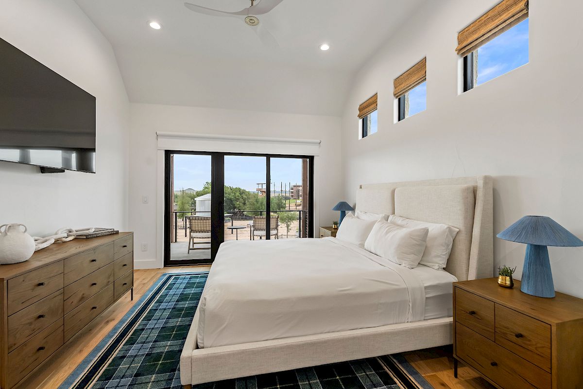 A bright, airy hotel-style bedroom with a king bed, matching nightstands, a wall-mounted TV, sliding door to a balcony, and a blue plaid rug.