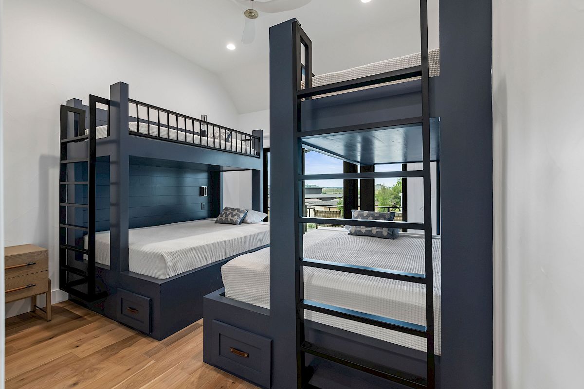A modern bunk bed room with two navy blue triple-level bunk frames, built-in drawers, a shared desk, and light wood flooring, bright and tidy.