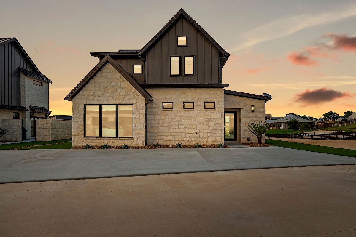 A modern two-story house with stone and dark wood siding, large windows, a spacious driveway, and a sunset sky in the background.