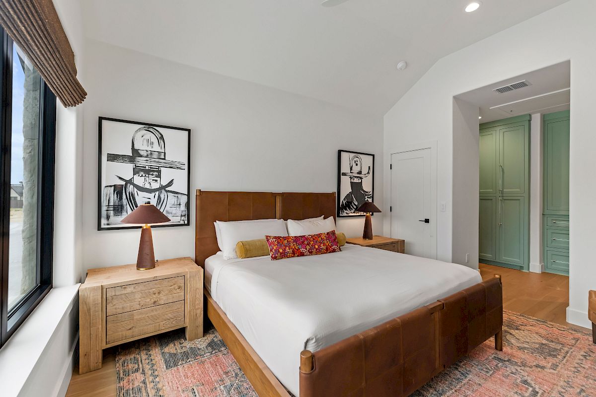 A stylish bedroom with a wooden bed, two nightstands, framed black-and-white art, a patterned rug, and a doorway leading to a mint-green hallway.
