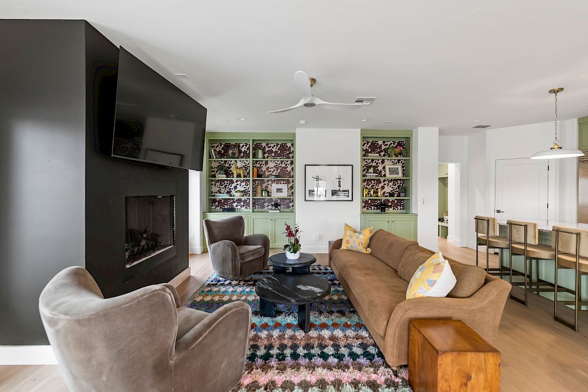 A modern, open-concept living room with a black accent wall fireplace, gray chairs, a brown sofa, colorful rug, and a kitchen/dining area in the background.