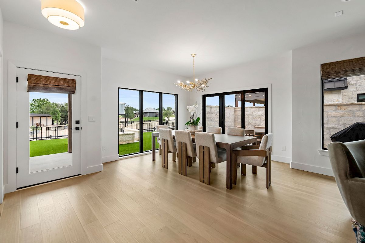 A bright dining room with a long wooden table and beige chairs, large glass doors/windows to the yard, light wood flooring, and a modern chandelier.