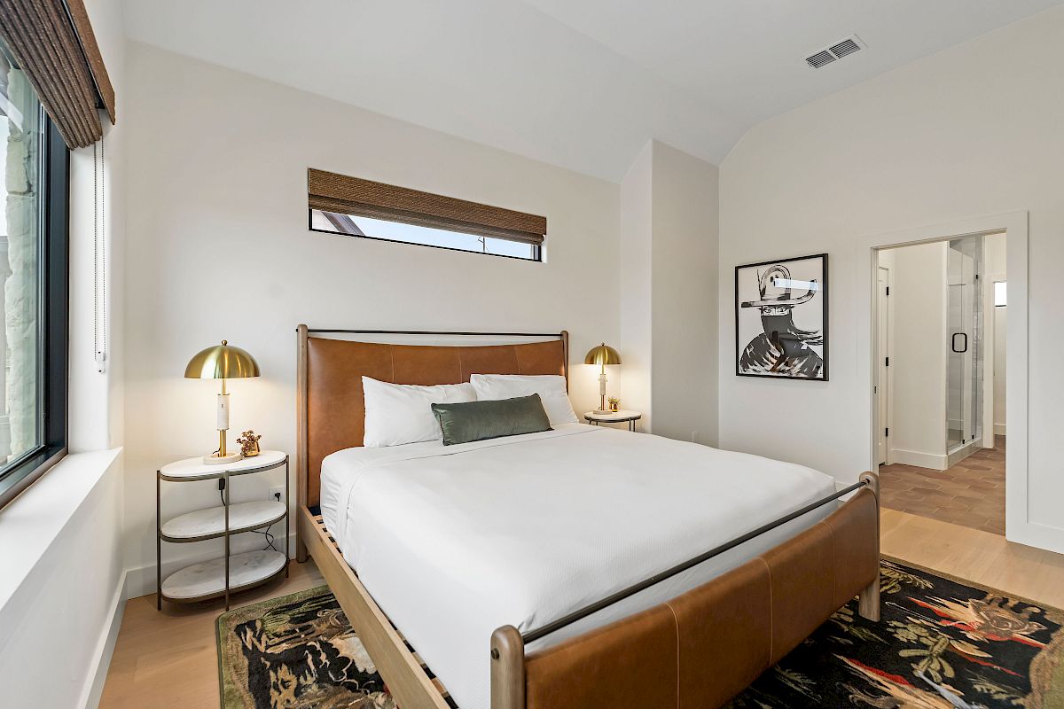 Cozy, modern hotel room with a wooden bed, white linens, two matching nightstands, lamps, a window, rug, and a framed art print near the doorway.