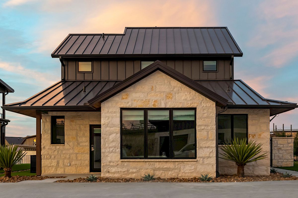 A modern stone-and-slate house with large windows and a metal roof in a desertscape, featuring a central bay window and small palms, at dusk.