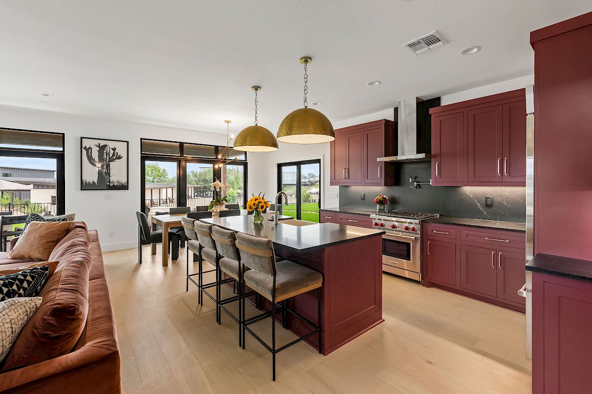 A modern open-concept kitchen with deep maroon cabinets, a large island with seating, stainless steel appliances, and a sunlit dining area.
