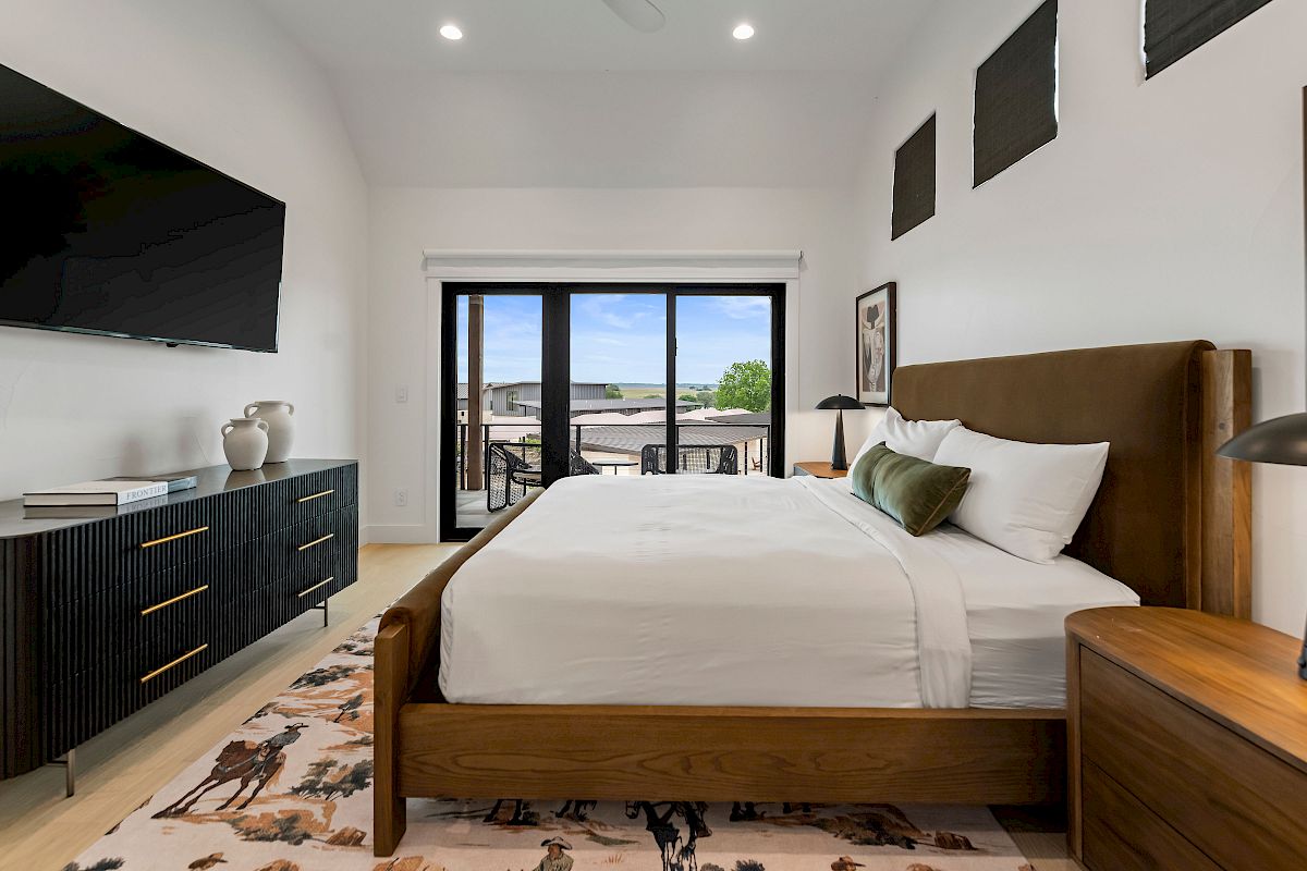 A modern bedroom with a wooden bed, white linens, and a green accent pillow; a dresser, wall-mounted TV, and a large sliding door to a balcony overlooking a sunny view.