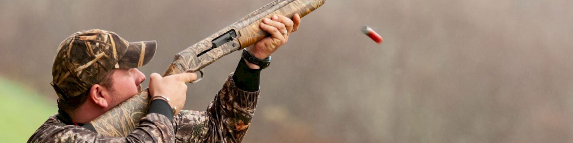 A hunter in camouflage aiming a shotgun, firing a shell mid-air on a grassy field.