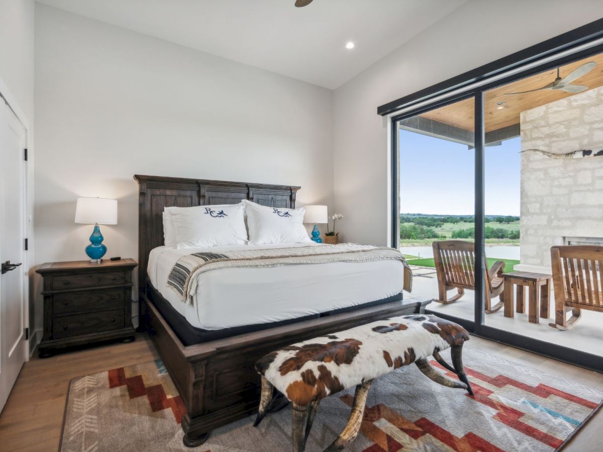 A bright, modern bedroom with a dark wood king bed, matching nightstands, a camel wool bench, and a colorful rug. Sliding glass doors open to a balcony with views.