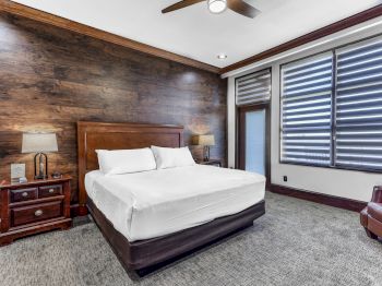A modern hotel room with a large wooden accent wall, king bed, two nightstands, lamps, a ceiling fan, and a window with striped blinds.