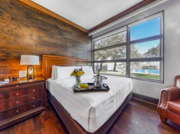 A cozy hotel room with a king bed, wooden accent wall, bedside drawers, a lamp, a chair, and a large window showing trees and a distant view, bright and inviting.
