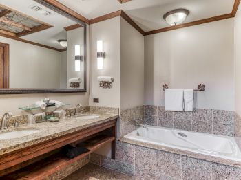 A luxurious bathroom with a double-sink granite vanity, large mirror, wall sconces, and a tiled soaking tub with a towel rack. Finish is warm wood and neutral tones.