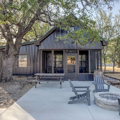 A quaint cabin with a screened porch is surrounded by trees. The outdoor area has Adirondack chairs arranged around a stone fire pit.