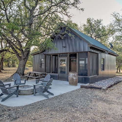 A small cabin with a metal exterior is surrounded by trees. It has a patio area with several chairs around a fire pit, under a clear sky.