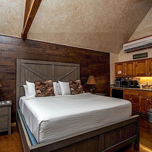 A cozy bedroom with a wooden bed, bedside tables, lamps, and a kitchenette equipped with appliances and cabinets, all in a rustic style.