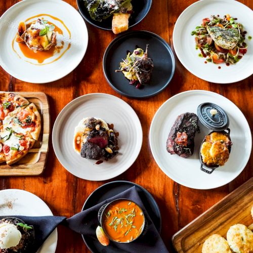 An assortment of plated dishes on a wooden table, including meats, pizza, soup, and biscuits.