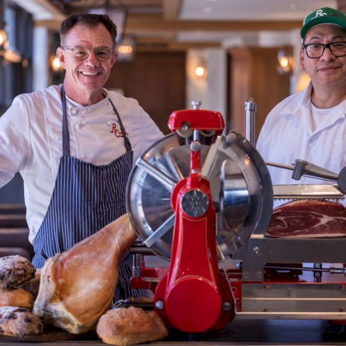 Two chefs stand beside a meat slicer and assorted bread and meats on a table.