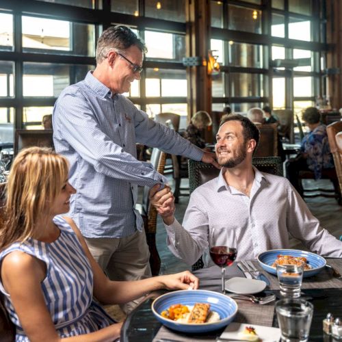 Three people are in a restaurant; two are seated with meals, and one is standing, shaking hands with the seated man, while the woman looks on.