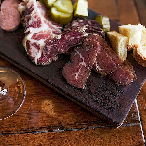 A charcuterie board with meats, pickles, and bread sits on a wooden table next to a glass of wine.