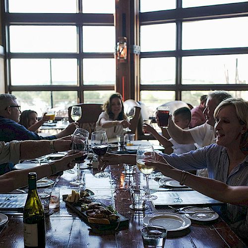 People sitting around a long dining table in a restaurant, raising their glasses to toast, with wine and food on the table, smiling.