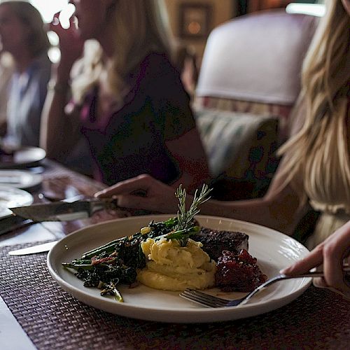 A person is dining with others, eating a meal with mashed potatoes, greens, and meat, paired with a glass of red wine, smiling while cutting food.