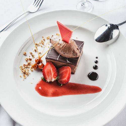 A chocolate dessert with strawberries, sauce, and a decorative element on a white plate, accompanied by a fork and spoon.