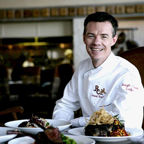 A chef in a white uniform is smiling and sitting at a table with various dishes in front of him.