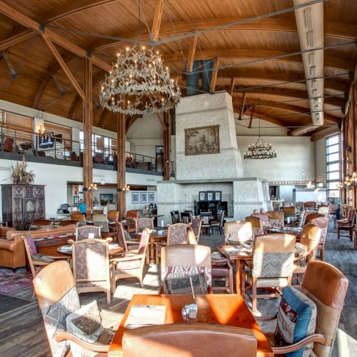 The image shows a spacious, rustic restaurant with wooden beams, chandeliers, and a large stone fireplace, filled with tables and chairs.