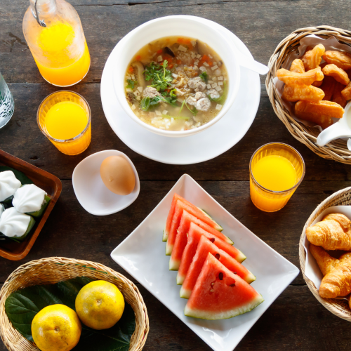 A breakfast spread with soup, watermelon, croissants, pastries, steamed buns, juice, water, and fruit on a wooden table.