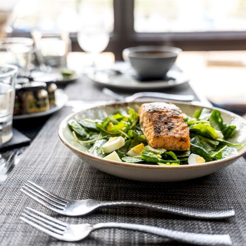 A grilled salmon fillet on a bed of spinach and vegetables, with dining cutlery on a table set for a meal by a window.