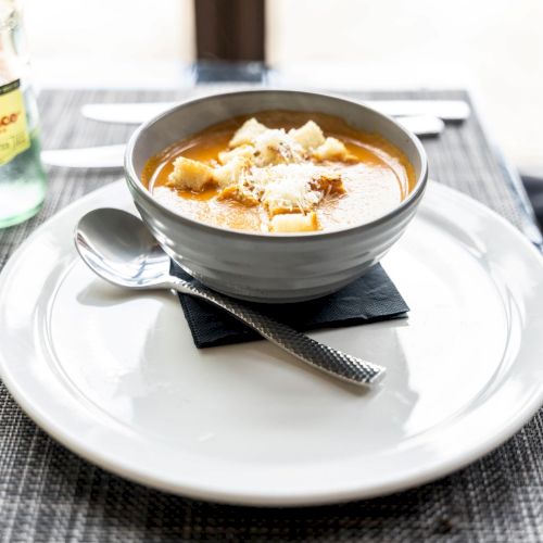 A bowl of soup topped with croutons on a white plate beside a spoon. There's a bottle of Topo Chico and a lime slice to the side.