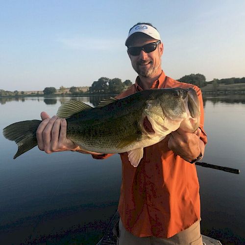 A person in an orange shirt and sunglasses is holding a large fish in front of a calm lake with trees in the background.