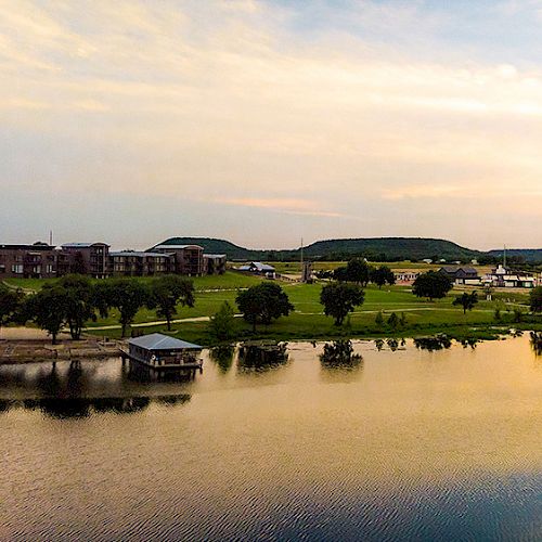 The image shows a serene lakeside with a small dock, several buildings in the background, and a picturesque sunset sky reflecting on the water.