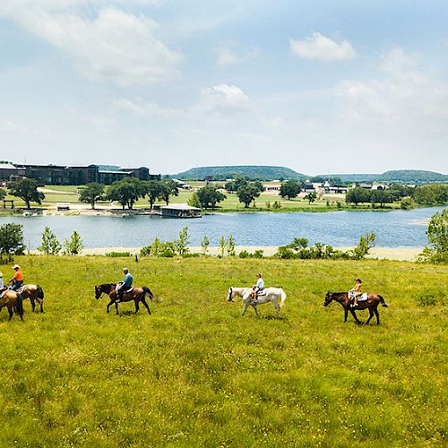 People riding horses in a grassy field with a lake and buildings in the background under a partly cloudy sky.