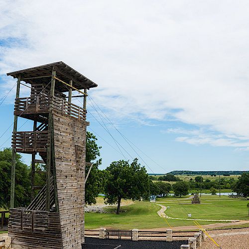 The image shows a wooden climbing tower with surrounding greenery and a small body of water in the background under a partly cloudy sky.