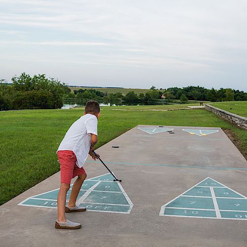 A person is playing shuffleboard outdoors on a concrete court with marked triangular scoring zones, surrounded by grassy fields and trees.