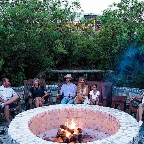 People are gathered around an outdoor fire pit, sitting on chairs and enjoying a peaceful evening in a lush garden setting.