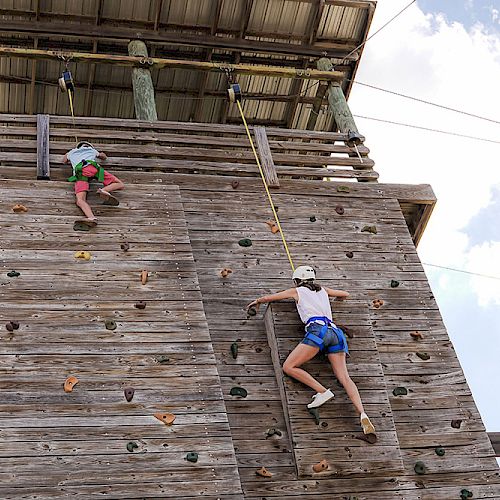 Two people are rock climbing on a wooden outdoor climbing wall while wearing helmets and harnesses under a partly cloudy sky.