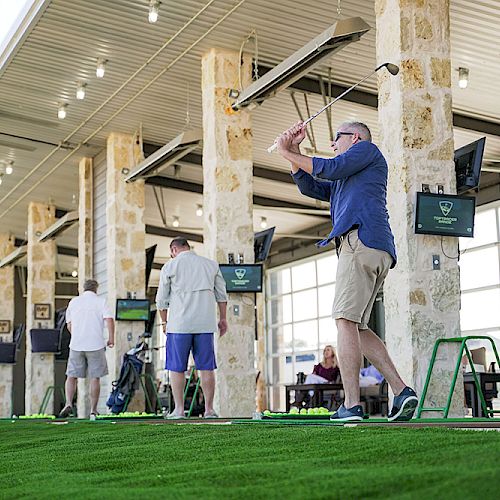 People playing golf at an indoor driving range. One man is swinging a golf club, while others are watching or preparing to play.