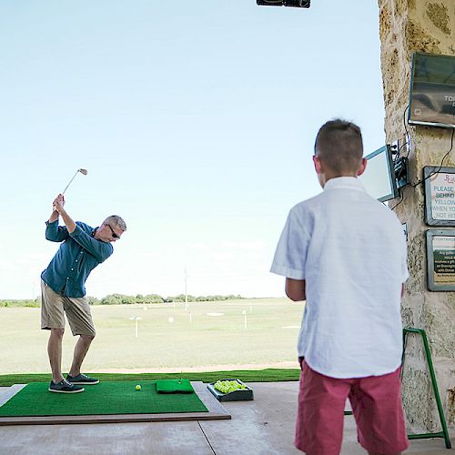 A person is practicing golf at a driving range while another looks on. There are signs and equipment visible, with a green field in the background.