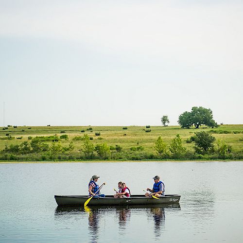 Three people are sitting in a canoe on a calm lake, surrounded by a green landscape and clear skies.