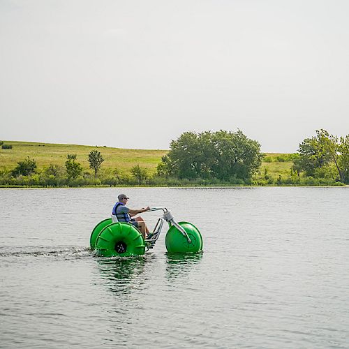 A person is riding a green water tricycle on a calm body of water with a grassy landscape in the background.