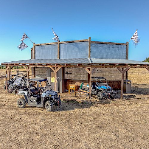 A rustic outdoor shelter with checkered flags, shaded seating, and several all-terrain vehicles parked in front on a dry open field, under clear skies.