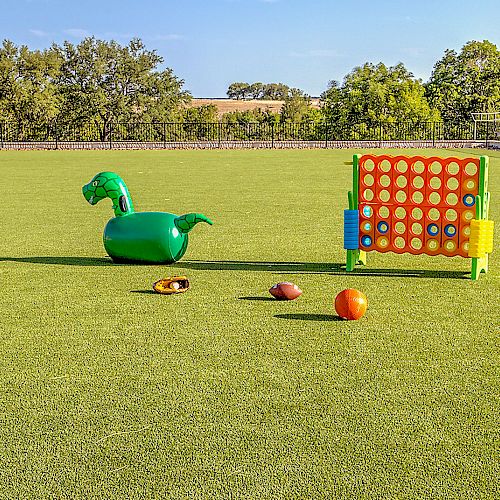 An outdoor play area with a large green field, an inflatable green dragon, a giant Connect Four game, a couple of balls, and some scattered toys.