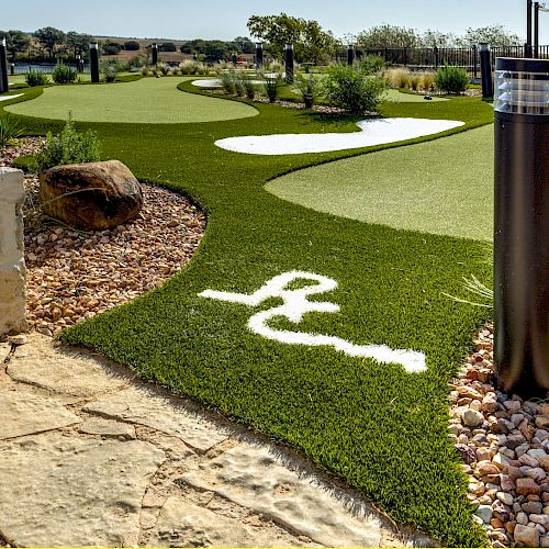 This image shows a landscaped mini-golf course with artificial turf, stone pathways, plants, and modern light fixtures. There is a white logo on the turf.