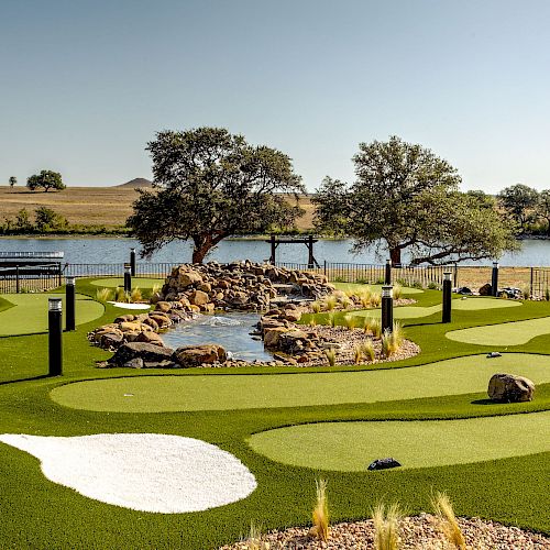 The image shows a landscaped mini-golf course with artificial turf, a small pond, and trees. In the background, there's a lake and an open field.