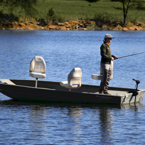 A person is fishing on a small boat in the middle of a lake, surrounded by calm water and natural scenery, with two empty chairs on the boat.