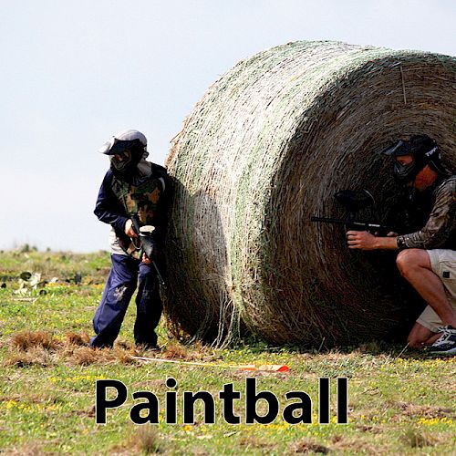 Two people are playing paintball, taking cover behind large hay bales. They are in a field with greenery, wearing paintball gear and masks.